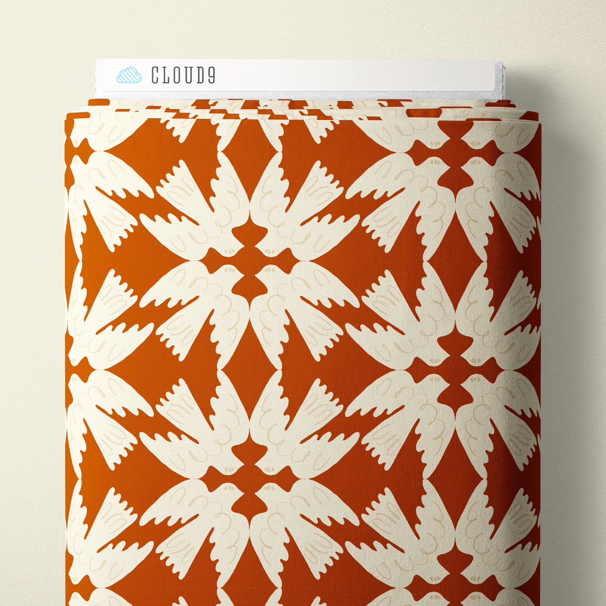A roll of patterned fabric with a repeating white bird and starburst motif on a warm orange-brown background, shown against a light beige wall. A white label at the top reads CLOUD9.
