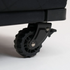 Close-up of a black caster wheel with a black plastic swivel bracket and a small silver metal screw, attached to a dark upholstered rolling bag on a white background.