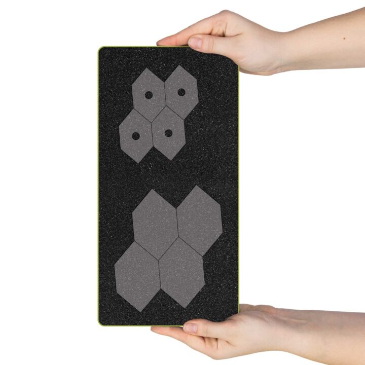 A black textured rectangular board with a thin yellow-green border, held by two hands against a white background. The board features two gray geometric hexagonal cluster designs, one smaller at the top and one larger at the bottom.