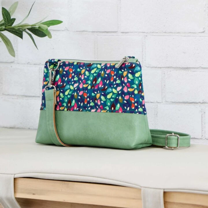 A color-blocked crossbody bag with a green faux-leather base and a colorful abstract patterned top panel, resting on a light-colored bench against a white brick wall.