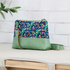 A color-blocked crossbody bag with a green faux-leather base and a colorful abstract patterned top panel, resting on a light-colored bench against a white brick wall.