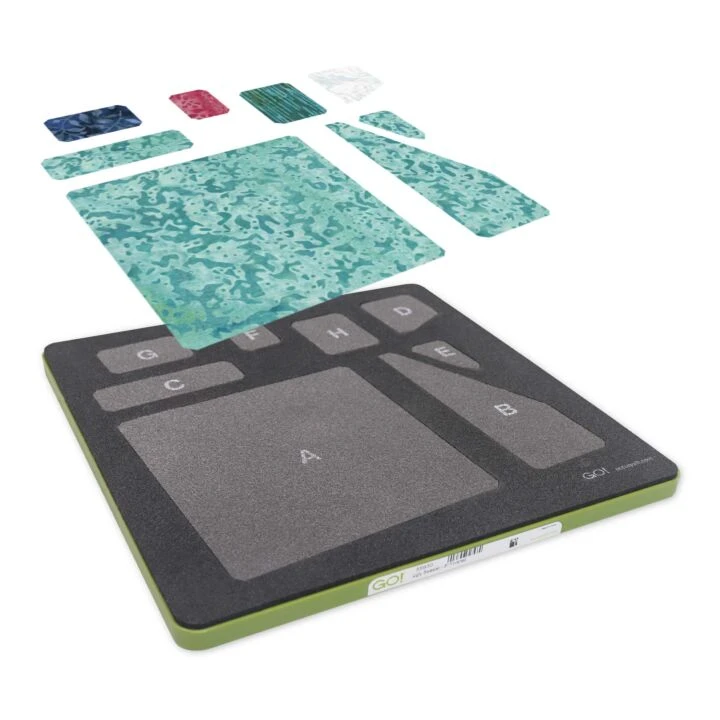 An AccuQuilt die cutting board with a black textured surface, olive green base, and labeled lettered zones (A, B, C, D, E, F, G, H), shown with fabric cuts floating above on a white background.