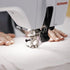 A close-up of a BERNINA sewing machine in white being operated, with a person's hands guiding white fabric under the needle.