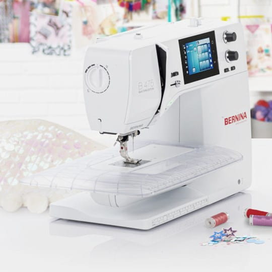 A white Bernina B-475 sewing machine with a digital screen and rotary dials, shown on a white surface with a blurred craft-themed background.
