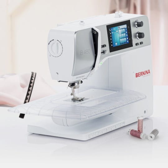 A white BERNINA sewing machine with a digital screen and control knobs, shown on a clean white surface with spools of thread nearby.