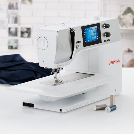 A white BERNINA sewing machine with a digital screen on a white table, with a dark navy fabric ball and thread spools in the foreground against a blurred white brick wall background.