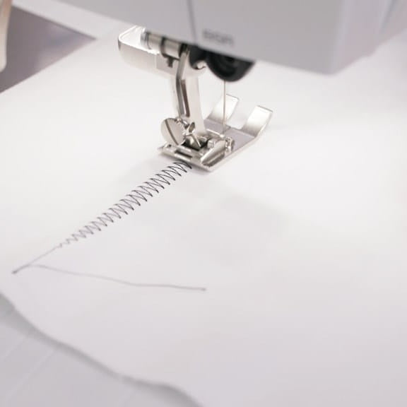 Close-up of a sewing machine presser foot stitching white fabric, showing a metallic silver presser and needle in action.