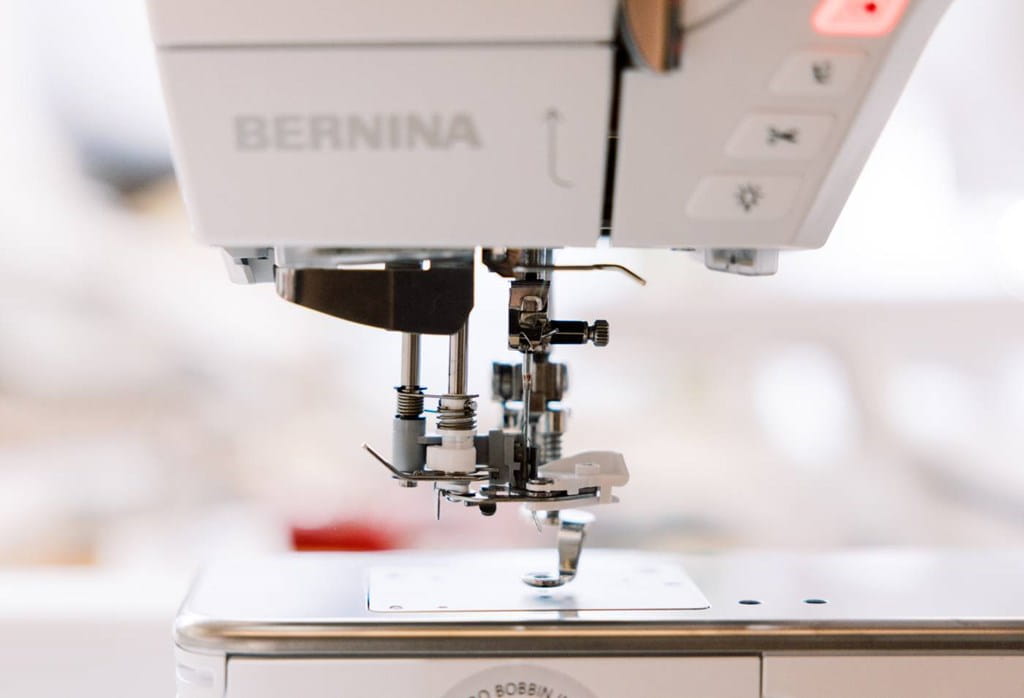 Close-up of a BERNINA sewing machine showing the needle area, presser foot, and part of the white machine body with a small red indicator light.