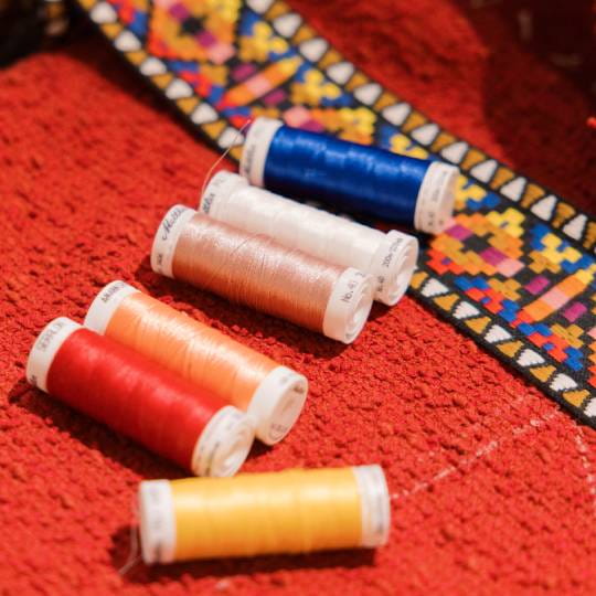 Five spools of embroidery thread in red, orange, white, blue, and yellow, arranged on a textured red surface next to a colorful embroidered pattern.