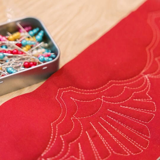 Red fabric with quilting design and a container of colorful pins on a wooden surface