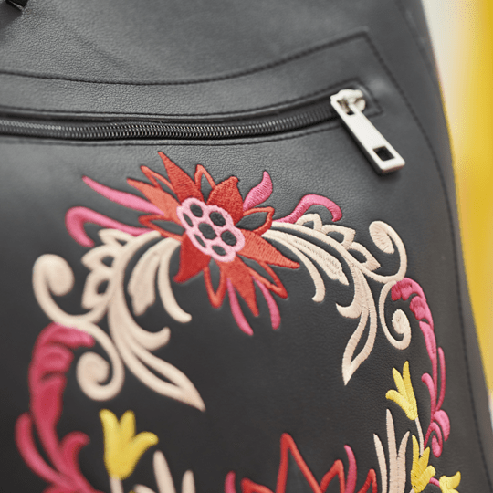 Close-up of a black leather or faux-leather bag featuring colorful floral embroidery in red, pink, cream, and yellow, with a silver zipper visible at the top.