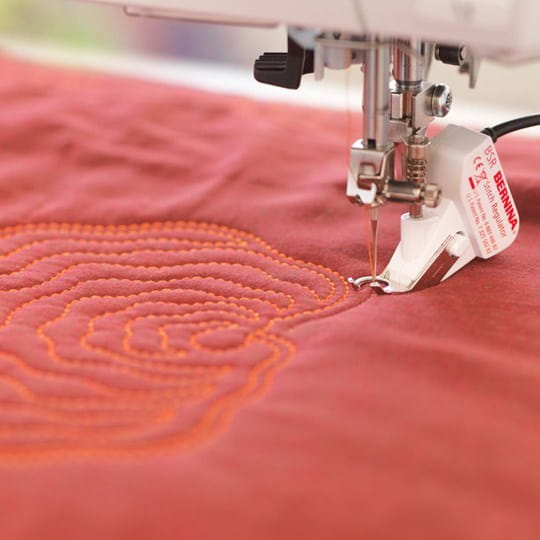 A close-up of a sewing machine needle stitching orange concentric floral patterns onto red fabric. The Bernina brand label is visible on the white machine component.