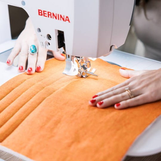 A close-up of a BERNINA sewing machine in use, stitching orange fabric with a person's hands guiding it. The BERNINA brand name is clearly visible on the white machine head.