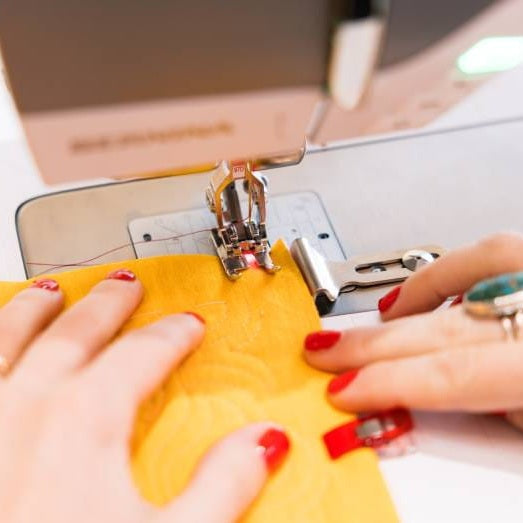 Close-up of a sewing machine in use with a person's hands guiding a bright yellow fabric under the press foot. The sewing machine is light gray, and the person has red-painted fingernails and a turquoise ring.