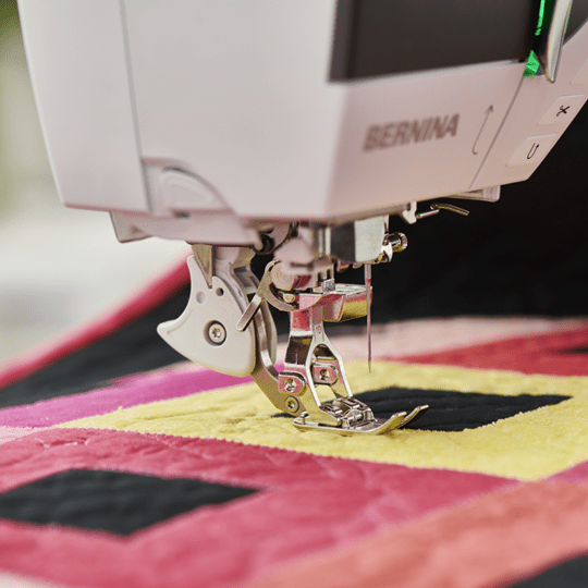 Close-up of a BERNINA sewing machine presser foot stitching a colorful quilt with pink, yellow, and black fabric blocks.