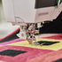 Close-up of a BERNINA sewing machine presser foot stitching a colorful quilt with pink, yellow, and black fabric blocks.