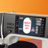A close-up of a modern sewing machine with a large touchscreen display showing the text 'CREATE WITH JOY' on the screen, set against an orange background.