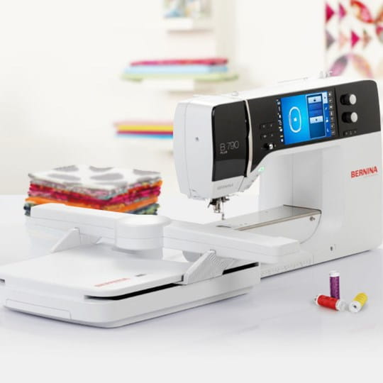 A white and black BERNINA sewing machine with a digital display screen, shown with its fabric feeddog attachment and spools of thread on a clean white surface.