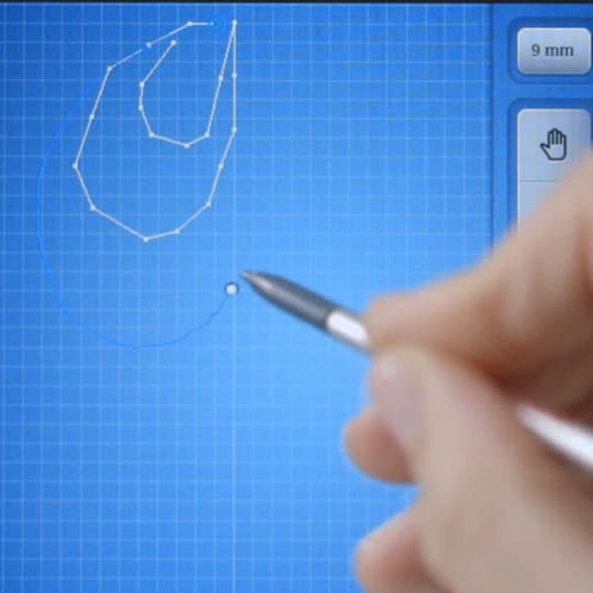 A close-up of a hand holding a silver stylus pointing toward a blue digital grid screen with a white outlined shape and interface buttons visible.
