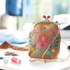 A small tan suede clutch purse with colorful floral embroidery, a gold-tone metal frame, and a gold chain strap, styled on a white table with sewing thread spools in a softly blurred background.