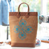 A tan canvas tote bag with brown leather handles and bottom trim, featuring a blue floral embroidered pattern on the front, set in a softly blurred sewing room environment.