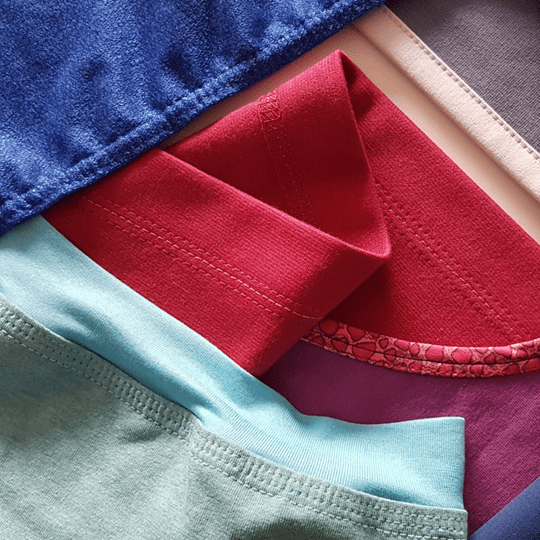 Close-up of folded cotton garments in several colors, including blue, red, pale blue, purple, and beige, showing fabric texture and stitching.