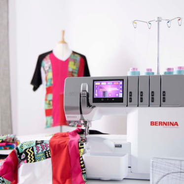 A BERNINA sewing machine in white and gray with a colorful digital display screen, shown in a bright studio setting with fabric and a patterned dress form in the background.