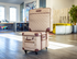 A quilted beige rolling suitcase with dark brown trim stands on a polished wood floor in a modern, sunlit room. A matching quilted storage bag is stacked on top of the suitcase.