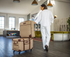 A woman in a white lounge outfit walks through a modern lounge area while pushing a stack of two quilted beige rolling suitcases with dark brown trim and wheels.