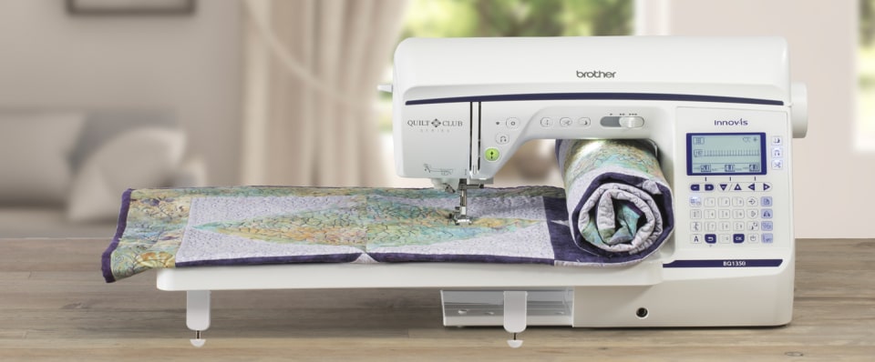 A Brother sewing machine on a wooden table with a quilt laid under the press head, shown in a softly blurred indoor setting.