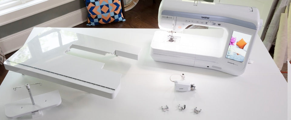 A white Brother sewing machine shown on a white table, with a matching white extension table beside it and small accessories placed in front. The room background includes a window and a patterned cushion.