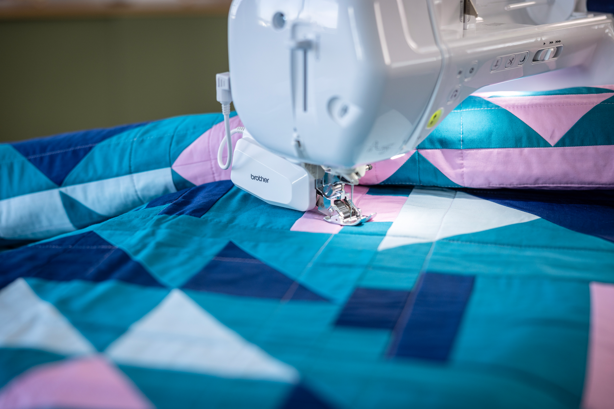 A Brother sewing machine in use, stitching a geometric quilt with teal, pink, and white triangular patterns.