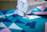 A Brother sewing machine in use, stitching a geometric quilt with teal, pink, and white triangular patterns.