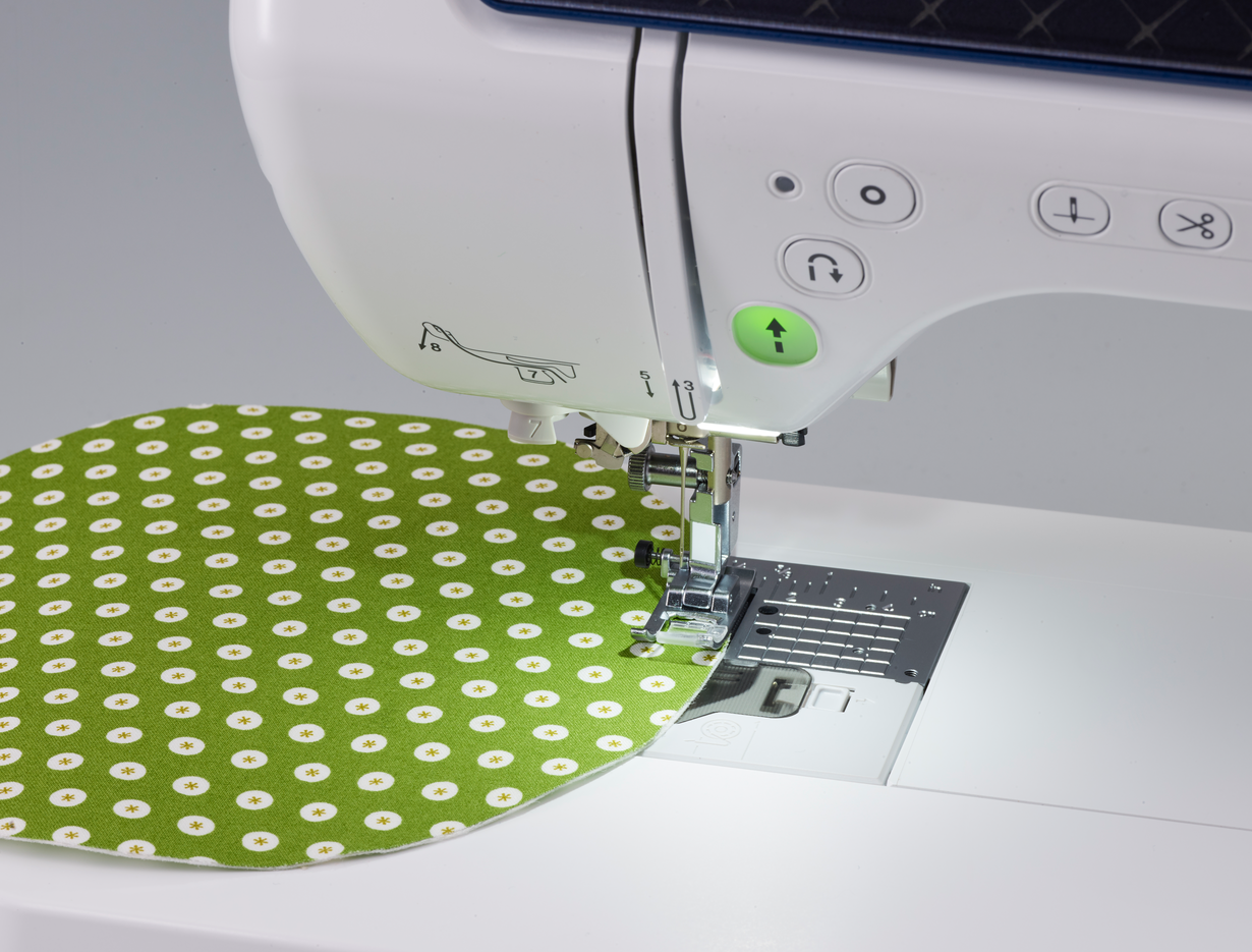 Close-up of a white sewing machine feeding a green fabric piece with white daisy patterns under the presser foot.