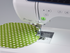 Close-up of a white sewing machine feeding a green fabric piece with white daisy patterns under the presser foot.