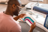 A man wearing glasses and a pink shirt uses a Brother embroidery machine to create a colorful embroidered design on a white fabric sheet.