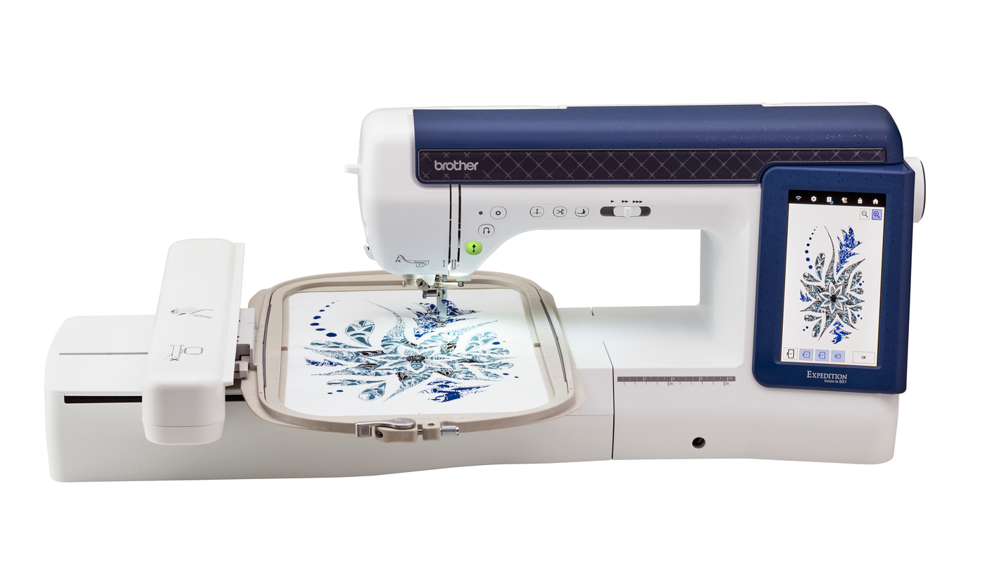 A Brother embroidery machine in white and navy blue, shown with an embroidery hoop containing a blue snowflake design on white fabric, against a solid white background.