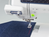 A close-up of a Brother sewing machine stitching a dark blue textured fabric, with the Brother logo visible on the machine body and a small green power button illuminated.