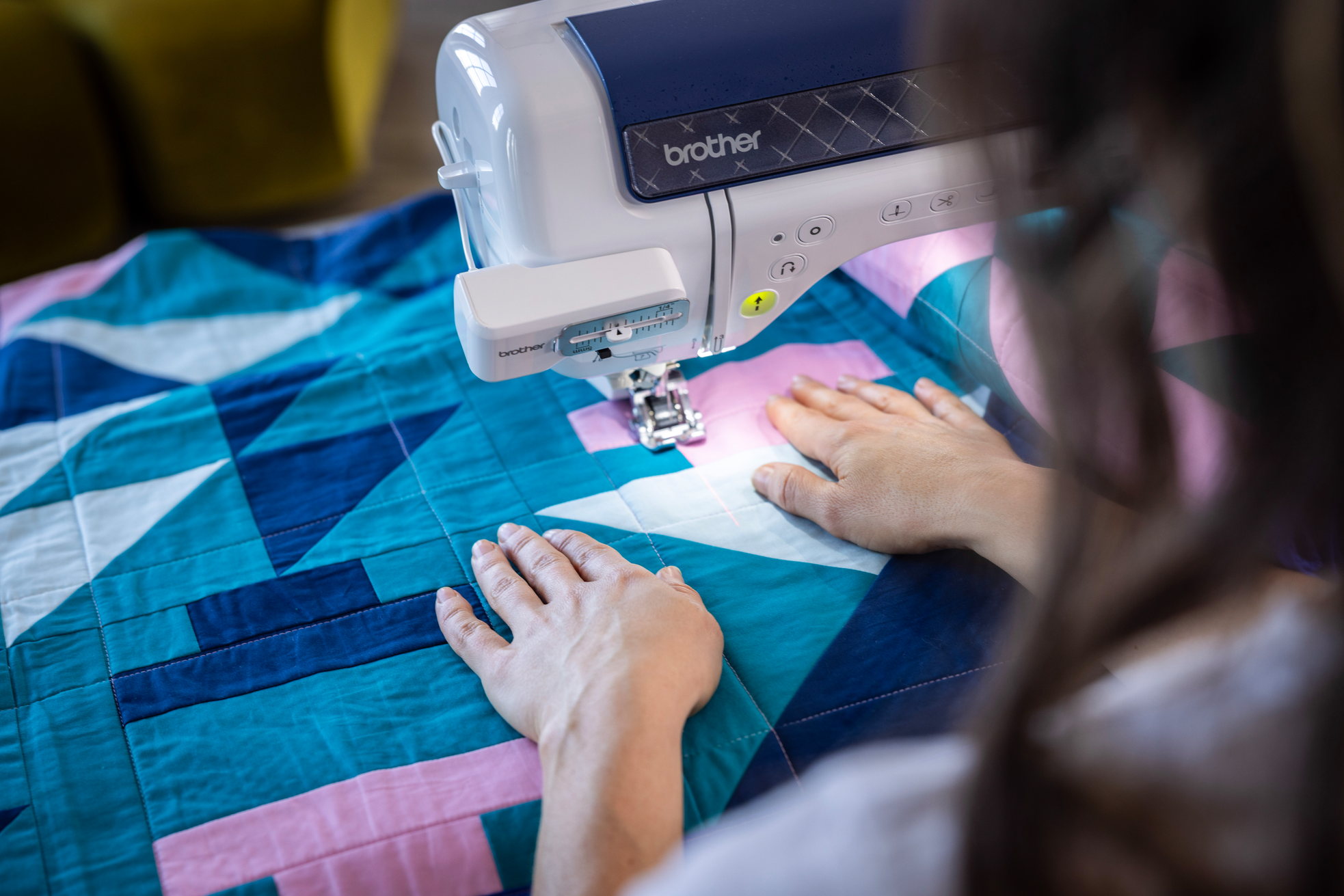 A Brother sewing machine in use with a person guiding a colorful patchwork quilt under the sewing head. The quilt features geometric patterns in teal, blue, pink, and white.
