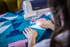 A Brother sewing machine in use with a person guiding a colorful patchwork quilt under the sewing head. The quilt features geometric patterns in teal, blue, pink, and white.