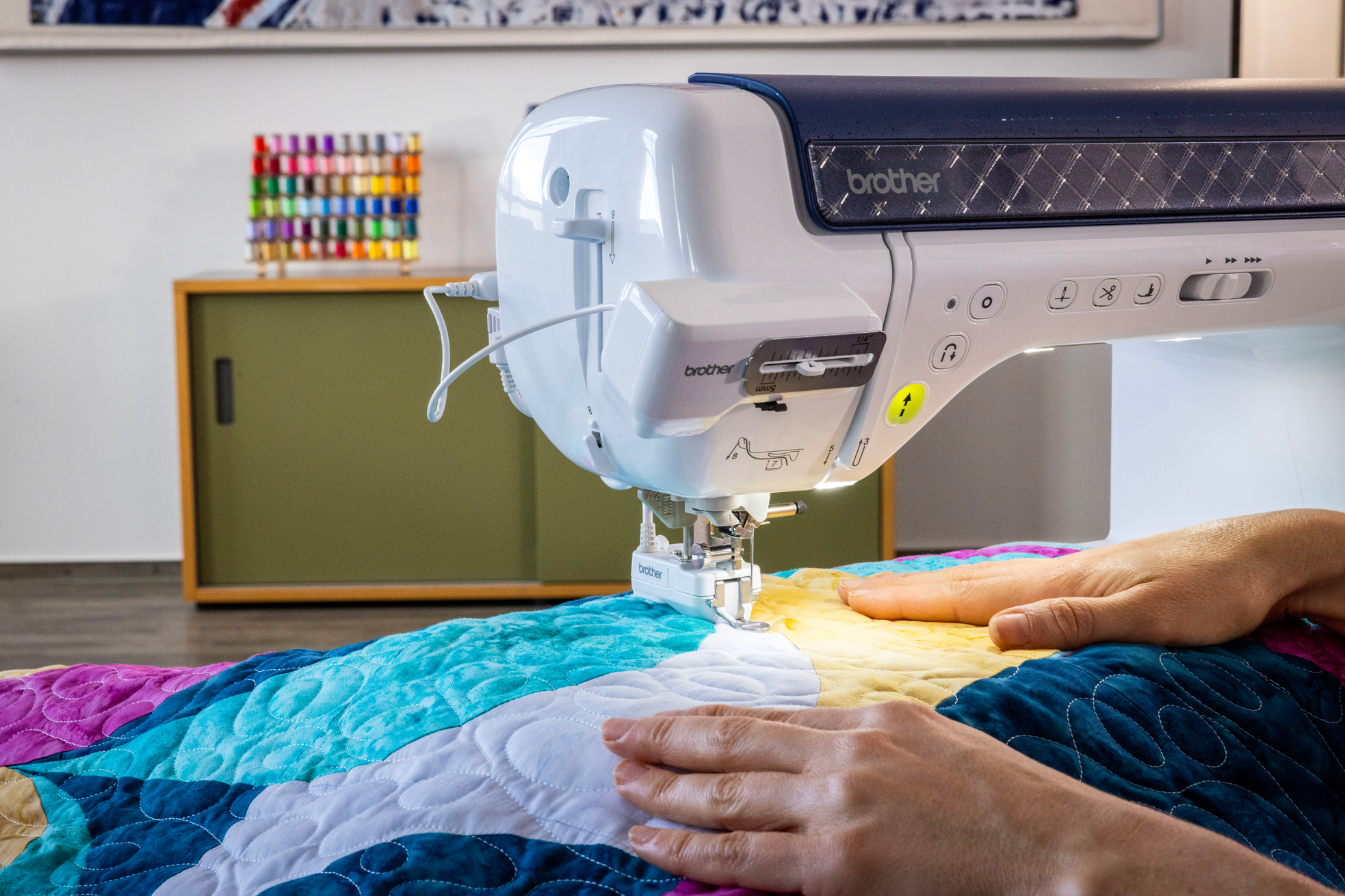 A Brother sewing machine in use, with a person's hands guiding a colorful quilt under the needle in a bright indoor room.