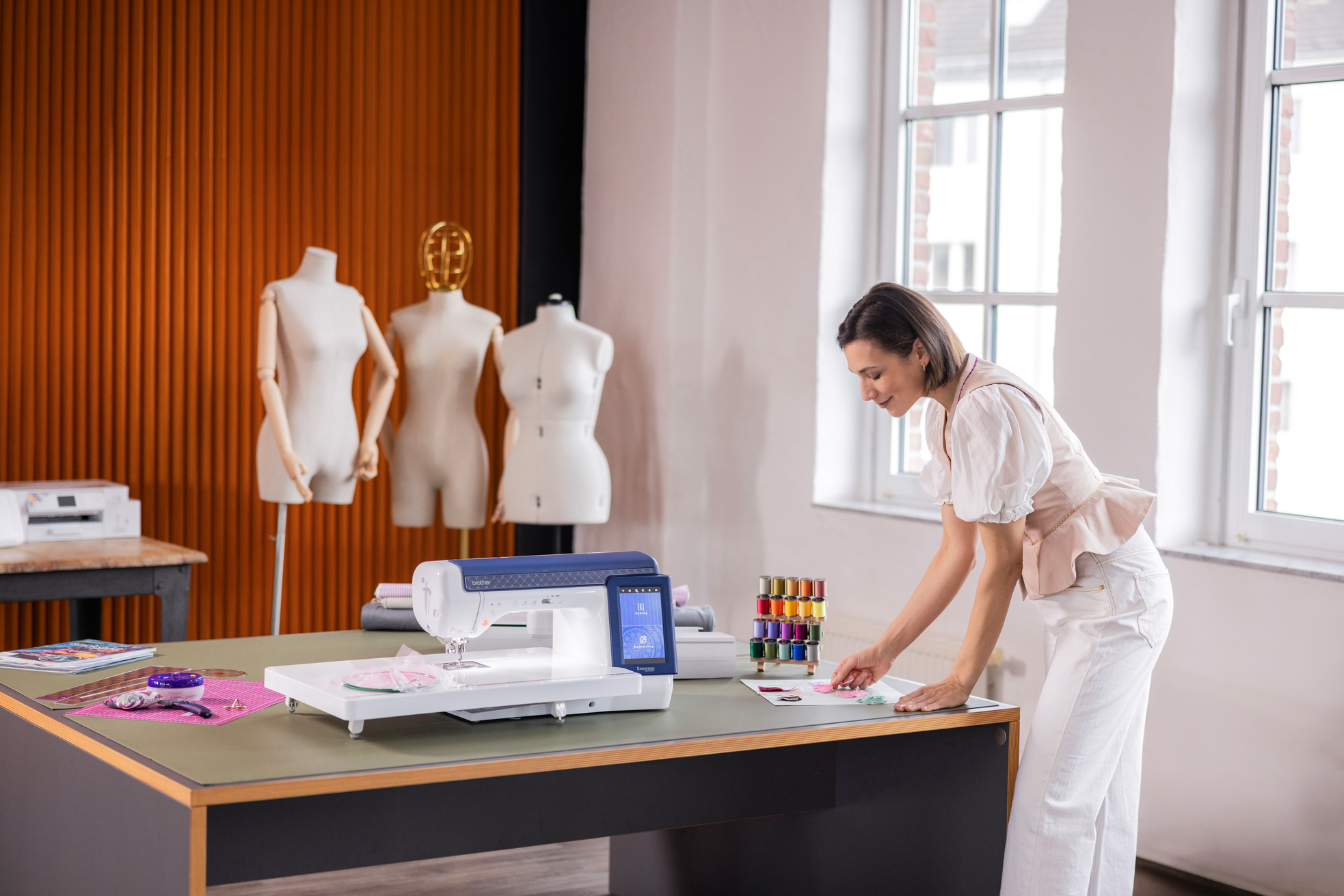 A woman in a white blouse and trousers stands at a sewing table in a bright room, working with fabric and a sewing machine. Three dress forms stand against an orange slatted wall in the background.