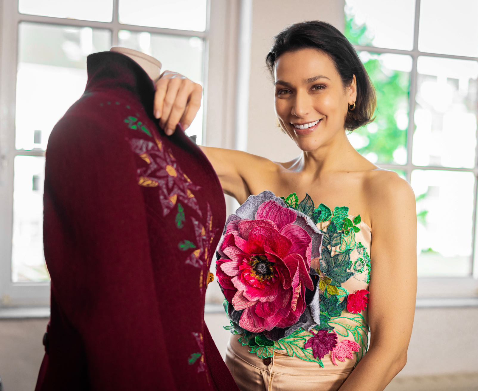 A smiling woman stands indoors beside a mannequin wearing a deep burgundy embroidered garment, while she holds a strapless top with large pink and green floral applique. Bright windows are in the background.