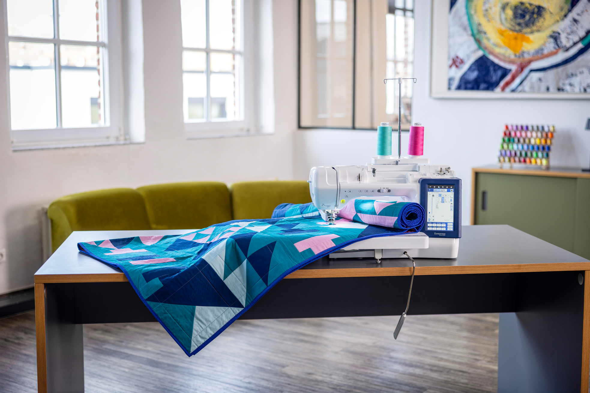 A sewing machine with colorful thread spools sits on a table next to a folded geometric quilt in blue, teal, and pink, set in a bright modern room with a green sofa and windows.