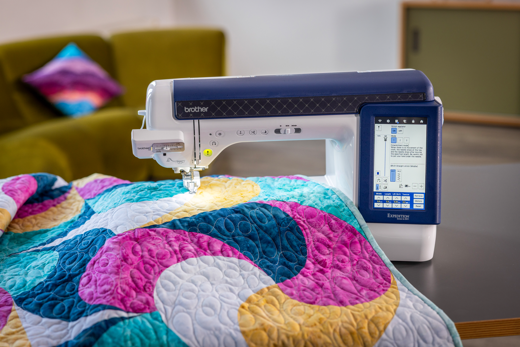 A Brother sewing machine with a digital touchscreen interface, shown quilting a vibrant, multicolored patchwork blanket in a cozy living room setting.