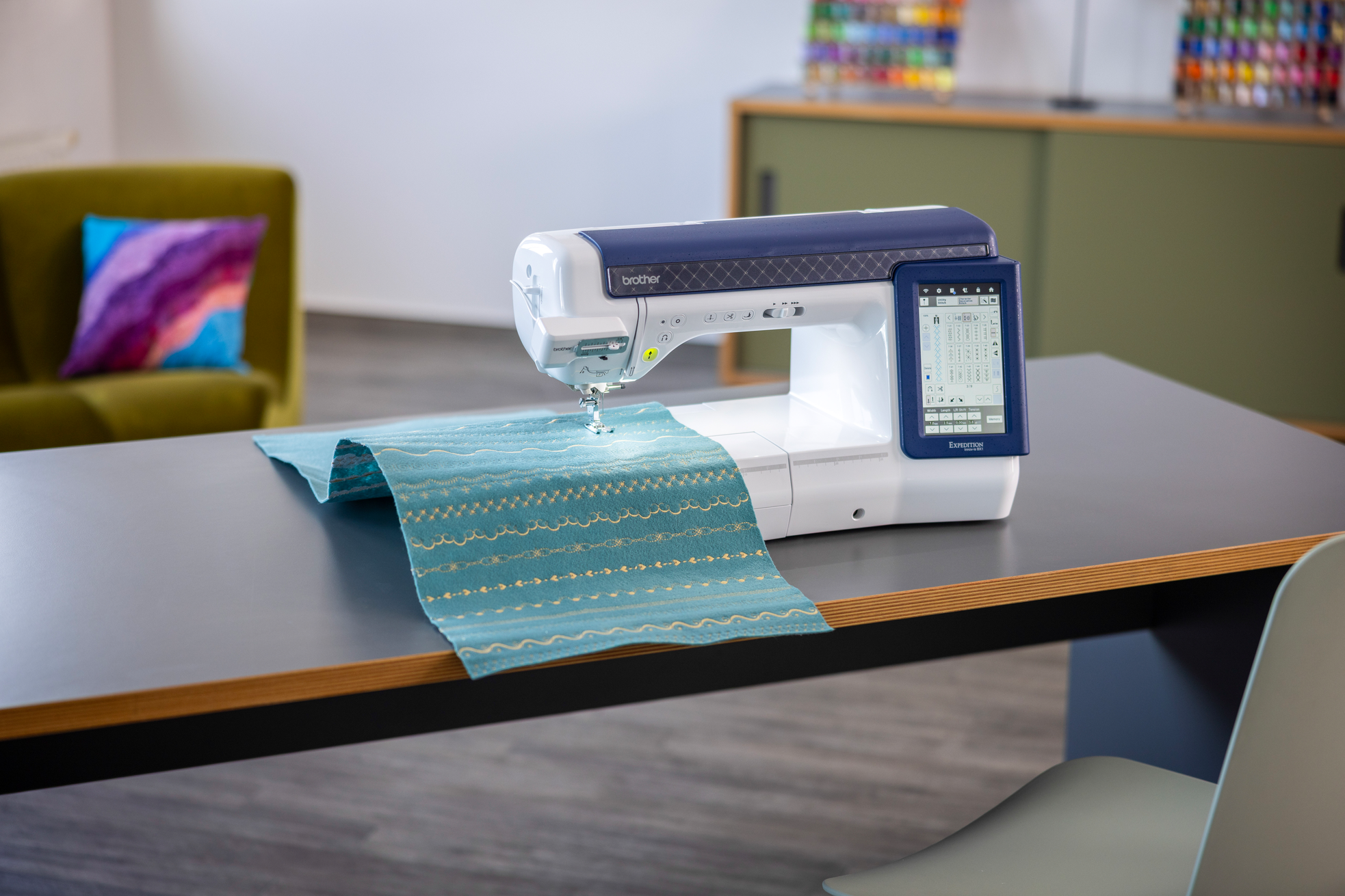 A Brother sewing machine sits on a dark table in a bright room, stitching a teal patterned fabric.