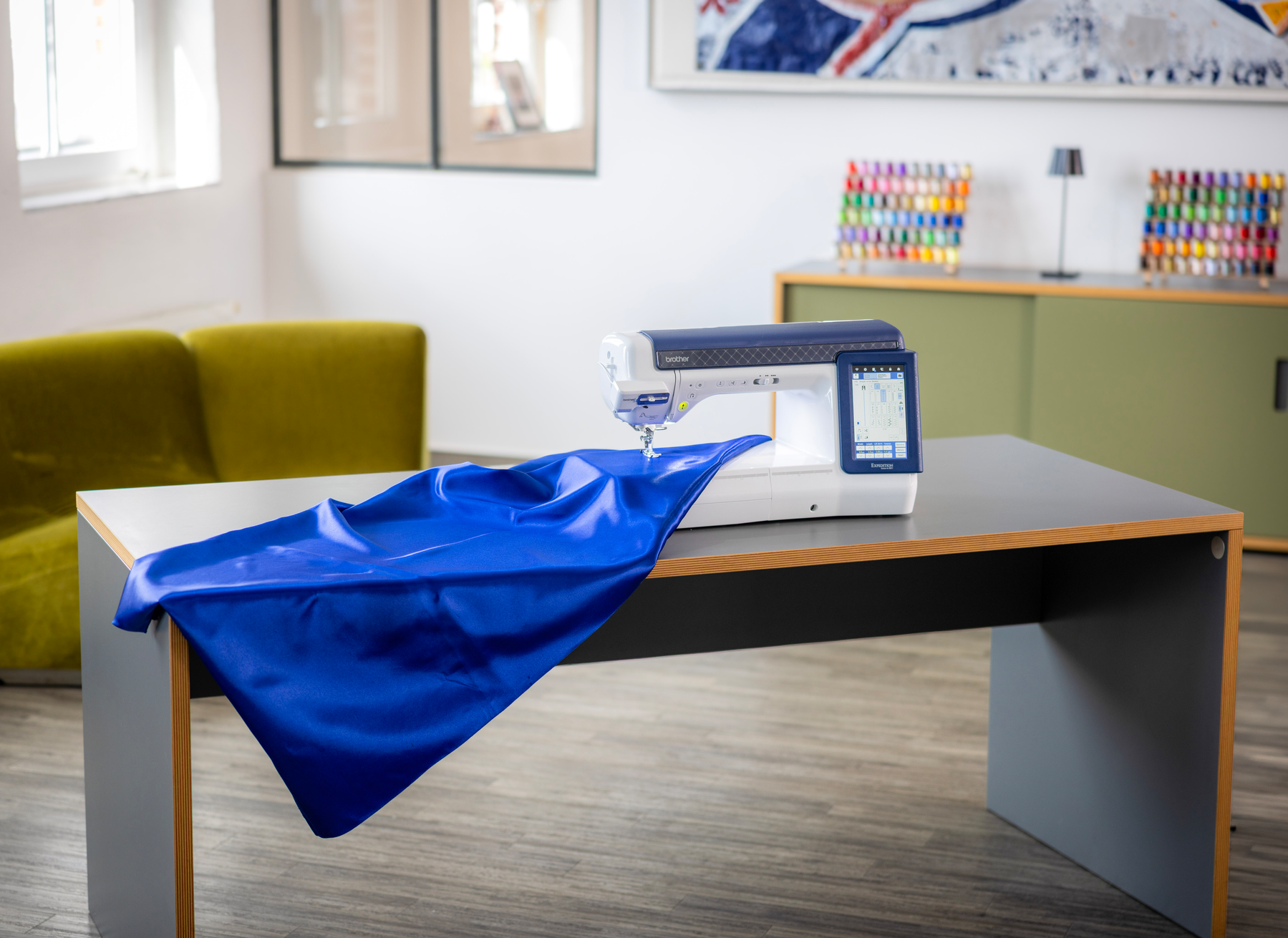 A white and blue sewing machine sits on a gray table in a modern room, with a bright blue fabric draped over the table in the foreground.