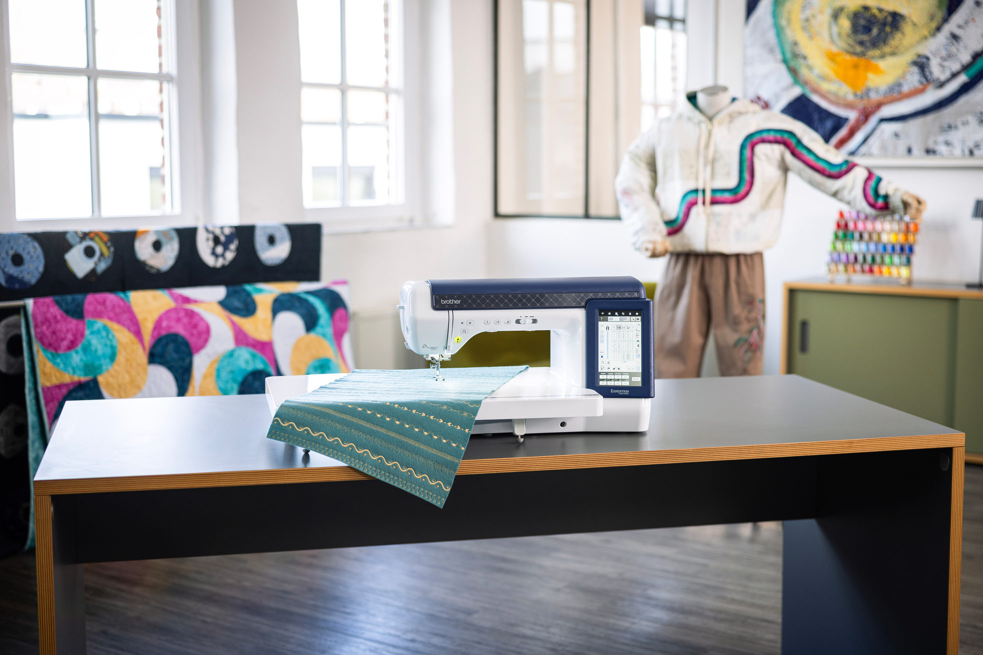 A white and navy sewing machine sits on a wooden table in a bright, modern craft room. A teal patterned fabric is draped over the machine, and a mannequin wearing a colorful quilted jacket stands in the background near a window.