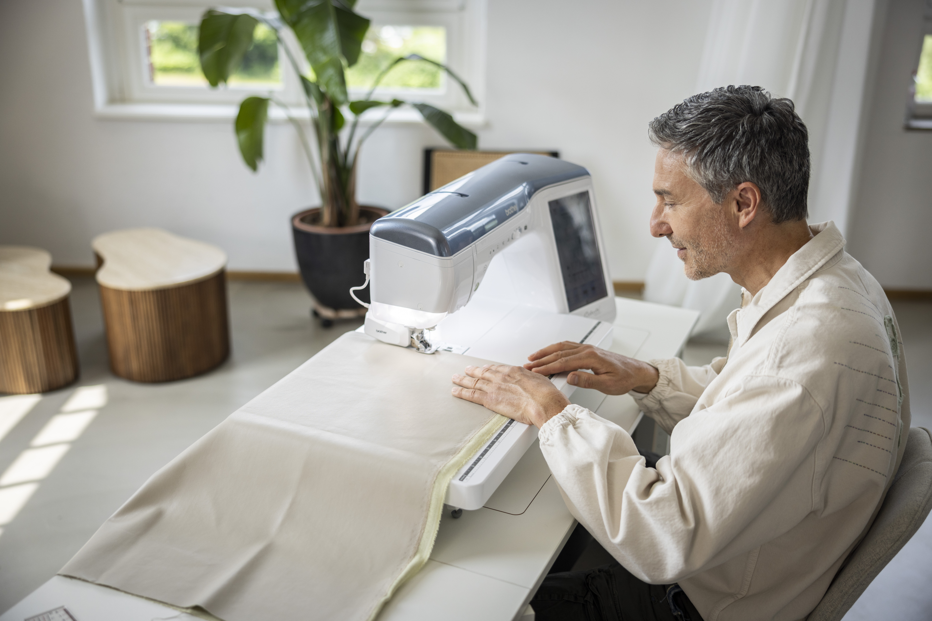 Man using a sewing machine in a bright room with plants and furniture.