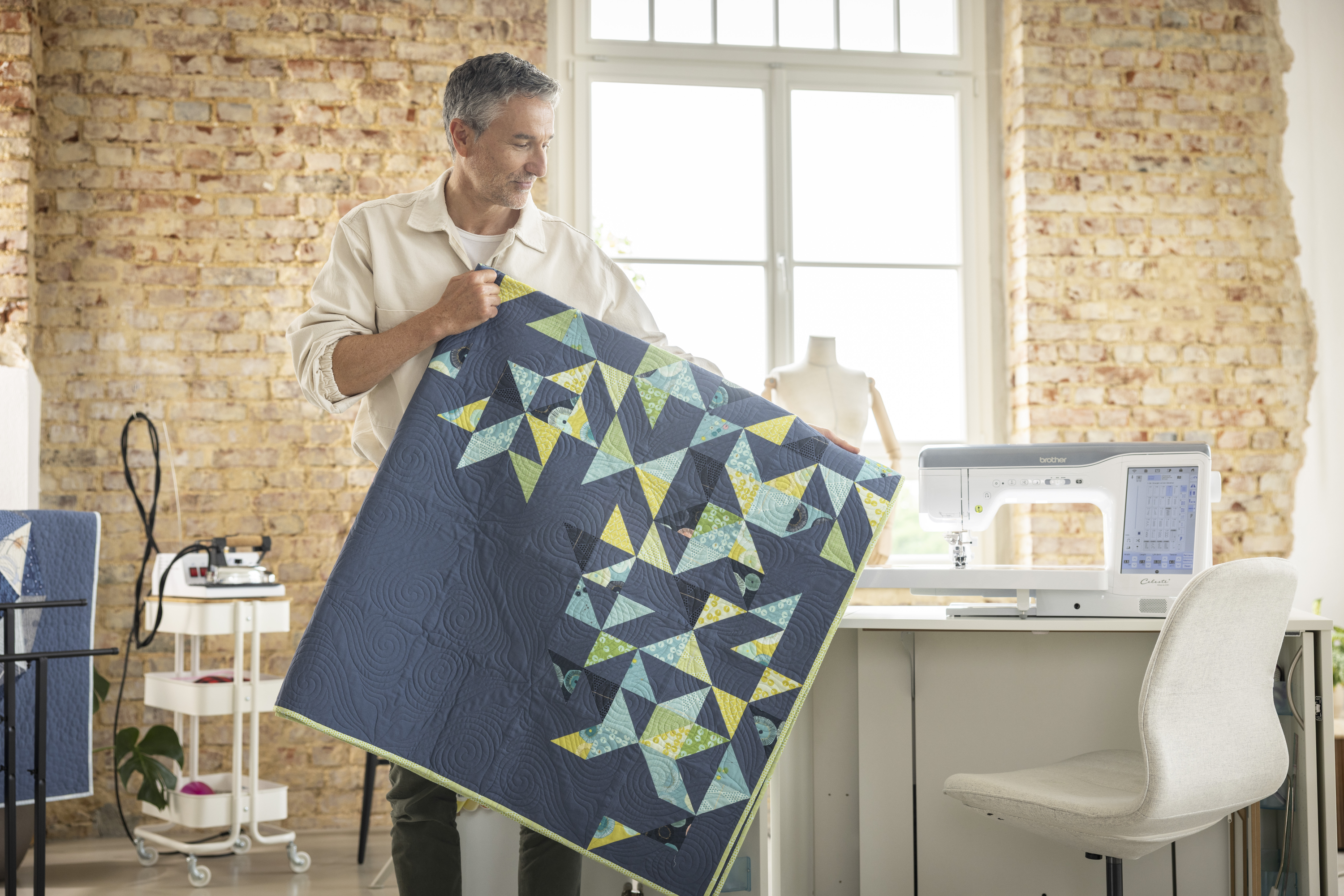 Man holding a geometric patterned quilt in a sewing room with a sewing machine and brick wall.